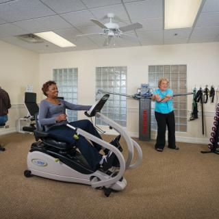 A group of people exercising indoors with fitness equipment, including a recumbent bike.