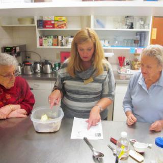 A group of three women, including two seniors, engage in a cooking session, following a recipe together in a kitchen.