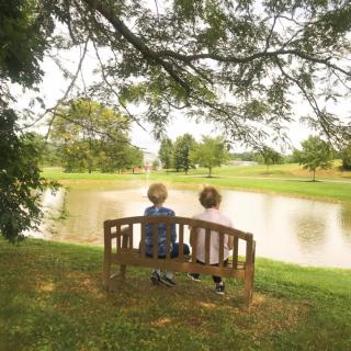 Two children sitting on a bench overlooking a lake in a peaceful park setting.