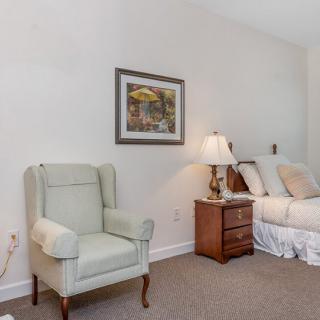 A cozy bedroom featuring an armchair, bed, bedside table, and a wall painting.