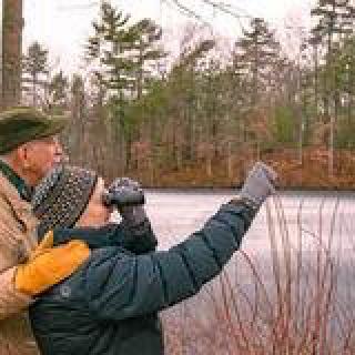 A couple is bird watching by a lake in a forest during winter.