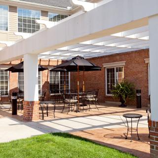 A sunny patio area with outdoor seating and umbrellas under a pergola, adjacent to a brick building.