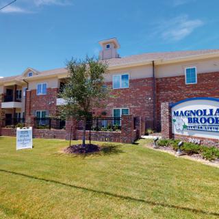 A brick apartment complex with a sign in front reading 'Magnolia Blooms' and well-maintained landscaping.