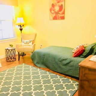 A cozy bedroom with a green bedspread, patterned rug, and a soft armchair near a lamp.