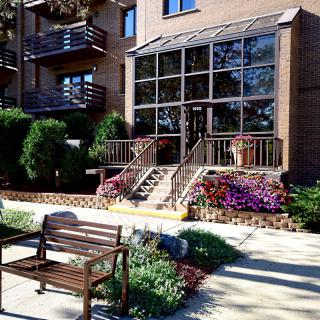 A modern apartment building entrance with a landscaped garden and bench.
