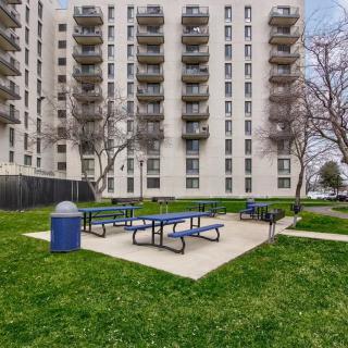 A residential apartment building with balconies overlooks a grassy outdoor area featuring picnic tables and a trash can.