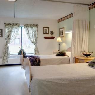 A well-lit hospital room featuring two twin beds with decorative elements such as curtains and a lamp.
