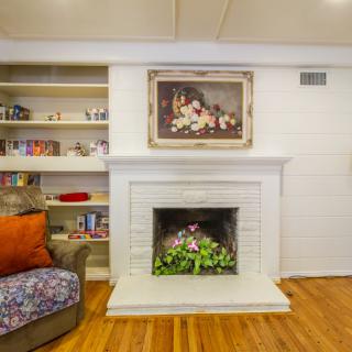 A cozy living room featuring a white fireplace with surrounding bookshelves, adorned with artwork and complemented by a comfy armchair and wooden flooring.