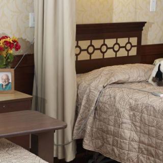 A cozy hospital room with two beds, adorned with quilts, separated by a curtain. A bedside table holds a framed photo and a vase of flowers.
