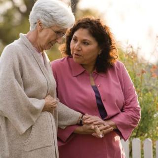A caregiver supports and converses with an elderly woman in a garden setting.