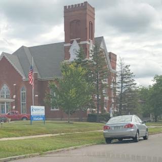A view of a red brick church with a tower, surrounded by trees and featuring a parked car on the street.