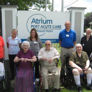 A group of elderly residents and healthcare staff posing together in front of Atrium Post Acute Care.