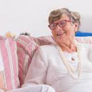 An elderly woman is sitting on a couch, smiling and wearing glasses.