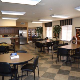 A spacious cafeteria with multiple tables and chairs, featuring a kitchen area and indoor plants.