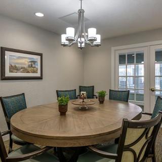 A cozy dining room with a round wooden table and upholstered chairs, illuminated by a modern chandelier. Small plants and artwork add to the decor beside elegant French doors.