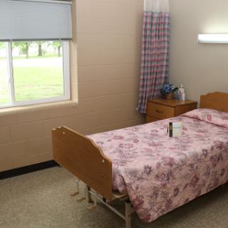 A simple hospital room featuring a bed with floral bedding, a window allowing natural light, and a nightstand with personal items.