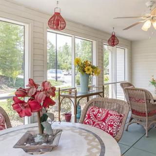 A bright sunroom with wicker furniture, red floral accents, and large windows letting in natural light.