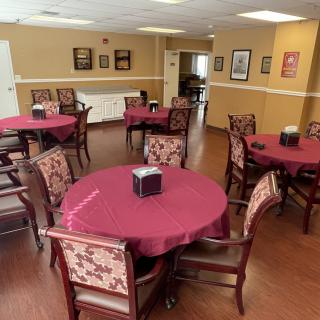 A neatly arranged dining area with round tables and upholstered chairs, each table covered with a maroon tablecloth and a napkin holder at the center.