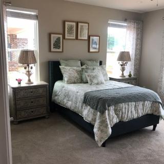 A cozy bedroom featuring a neatly made bed with patterned bedding and two nightstands topped with lamps. Framed artwork decorates the wall above the headboard, and light streams in through two windows.