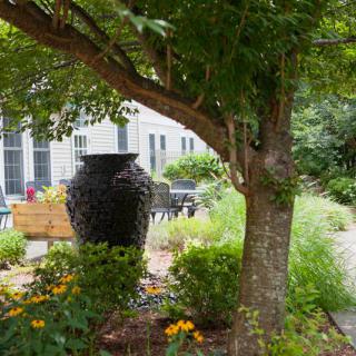 A peaceful garden scene with a large urn, colorful flowers, and lush greenery under a tree.