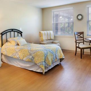 A cozy bedroom featuring a single bed with a patterned quilt, a chair and table set by the window, and bright natural lighting.