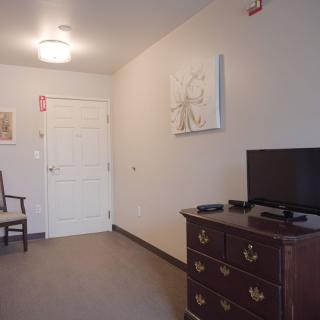 A simple room featuring a dresser with a television, a chair, and a door with wall art.