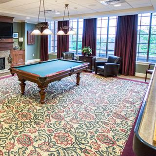 A well-lit billiards room featuring a pool table, elegant carpet, and comfortable seating by large windows.