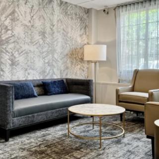 A modern living room featuring a dark gray sofa, an armchair, a round coffee table, and a tall floor lamp, set against a textured wall and large window with curtains.