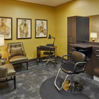 A modern salon interior featuring a styling chair, desk, artwork on the walls, and a small waiting area with comfortable seating.