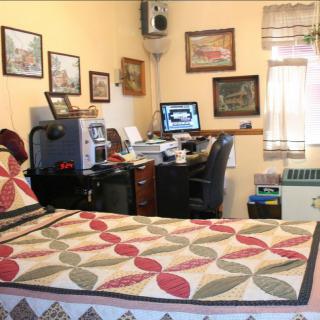 A cozy bedroom with a bed and an integrated home office setup, featuring a computer on the desk surrounded by framed pictures.