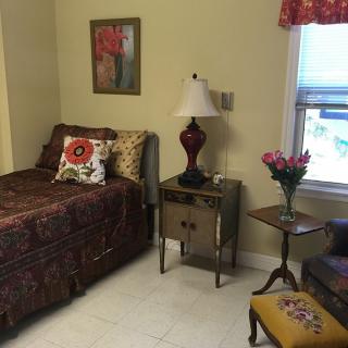 A cozy bedroom with a single bed, decorative pillows, and a bedside lamp next to a small table with flowers.
