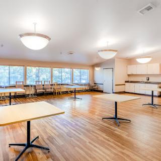 A spacious interior room with wooden flooring, featuring several tables and ample natural light from large windows.