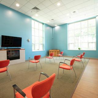 A bright and spacious meeting room with red chairs arranged in a circle, featuring a television and a children's play area.