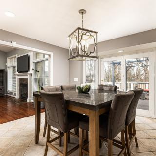 A modern dining room with a wooden table, upholstered chairs, and a decorative chandelier above, featuring large windows and hardwood flooring.
