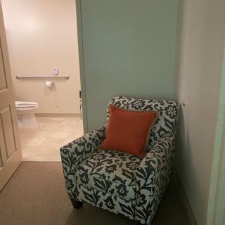 An armchair with a decorative pillow sits near a hallway leading to a bathroom.