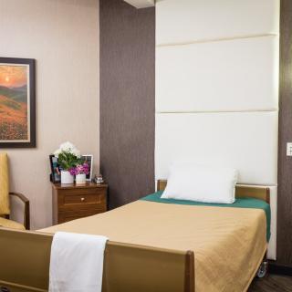 A tidy hospital room with a single bed, chair, and nightstand adorned with flowers.