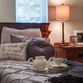 A cozy bedroom scene featuring a neatly made bed with decorative pillows, a tea set on a tray, and a softly lit lamp on the nightstand.