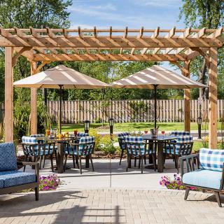 A cozy outdoor patio with comfortable seating under a wooden pergola, featuring umbrellas and surrounded by lush greenery.