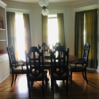 A cozy dining room with a wooden table surrounded by chairs, light filtering through curtains covering large windows.