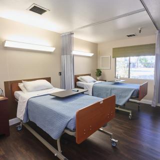 A hospital room with two neatly made beds, each equipped with a small table and situated in a brightly lit space.