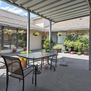 A covered patio area with a glass table and chairs, surrounded by potted plants and hanging baskets under sunny skies.
