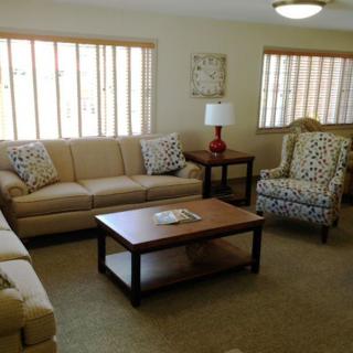 A cozy living room with sofas and armchairs arranged around a coffee table, illuminated by natural light from large windows.