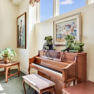 A cozy room featuring a piano with decorative sculptures on top, surrounded by artwork and plants, bathed in natural sunlight.