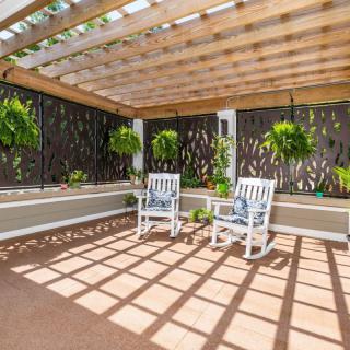 A serene patio area with a pergola providing shade over two white rocking chairs, surrounded by lush hanging plants.