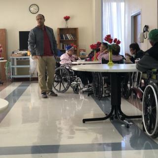 A caregiver interacts with elderly residents in wheelchairs inside a nursing home.