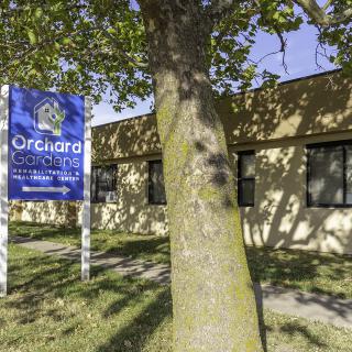A blue sign stands next to a tree in front of a beige building.