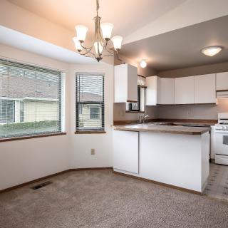 A modern kitchen with white cabinets and an open dining area, featuring a chandelier and large windows.