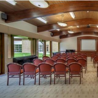 An empty conference room with rows of chairs facing a projection screen, featuring large windows and a carpeted floor.
