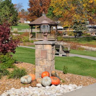 A scenic park view featuring a stone pillar adorned with pumpkins, set against the backdrop of a gazebo and vibrant autumn foliage.