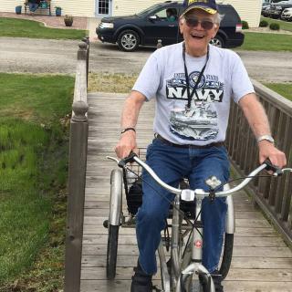 A person wearing a Navy t-shirt and hat, riding a bicycle over a wooden bridge.